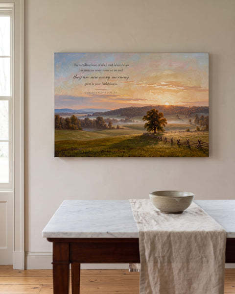 Painting-style sunrise meadow canvas with Lamentations 3:22-23 hanging on a greige plaster wall above a marble-topped console