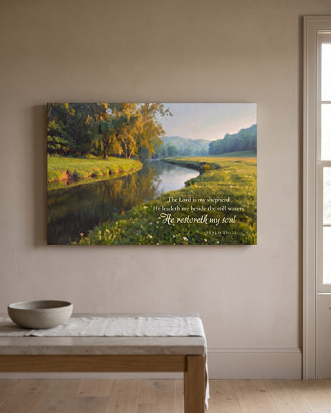 Painting-style canvas of a quiet stream winding through a green meadow with Psalm 23 lettering, hanging on a warm taupe plaster wall above a stone-topped console