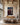 The largest 32 by 48 inch size on a tall plaster wall in a beamed great room, linen sofa and oak credenza below, stone chimney at right