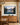 Sunrise over a mirror-still lake canvas above an oak sideboard with a bowl of green pears in a dining room