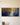 Extra-wide panoramic canvas of Jesus kneeling in prayer under a golden tree, centered on a warm cream plaster wall above a pale oak table