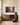 Woman kneeling by a walnut credenza with books in her lap, looking up at a painting-style Trust in the Lord forest canvas on a cream panelled wall