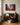 Woman with short silver hair kneeling by a walnut sideboard, unpacking a wooden nativity figure and looking up at the manger canvas