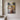 Vertical canvas of a lamb running through a stream toward Jesus kneeling with open arms, hung on a warm greige plaster wall