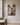 Vertical canvas of a lamb running through a stream toward Jesus kneeling with open arms, hung on a warm greige plaster wall